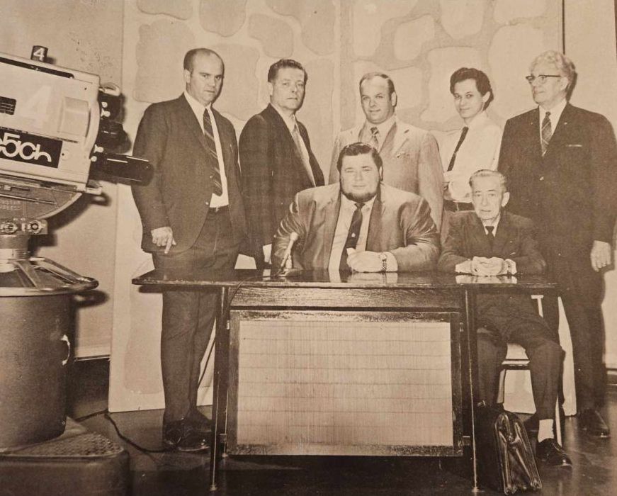 A behind the scenes shot of International Wrestling from a program includes, left to right standing, Ed Brown (producer), Larry Knoke (program manager), Al Zinck (promoter), George Flie (scriptwriter), and Curly Aguire (timekeeper), with George Cannon (commentator) and Johnny Wall (ring announcer) seated.